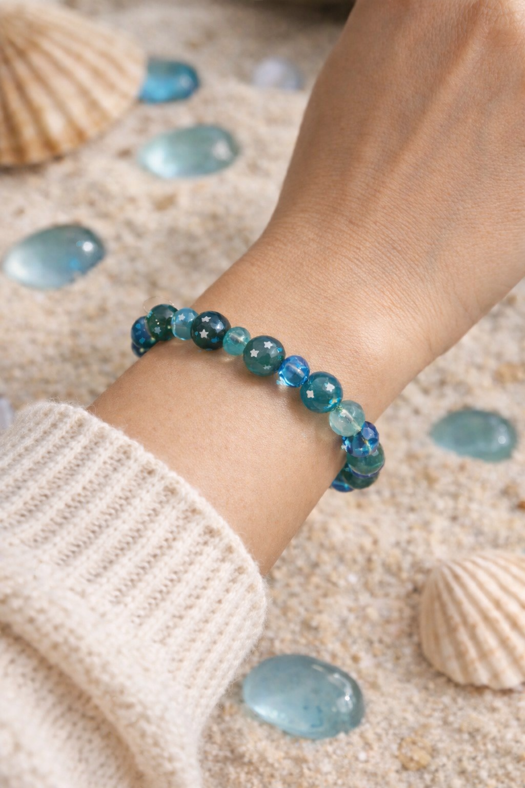 Coastal-inspired flat lay of a blue and teal beaded star bracelet styled with seashells and sea glass on sand