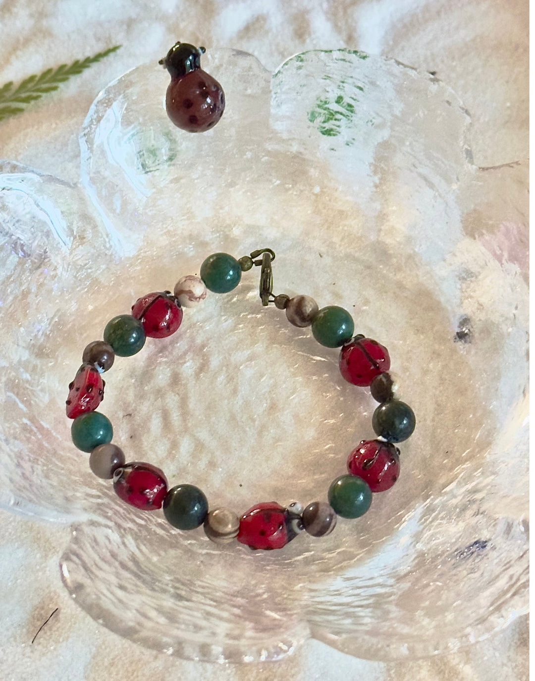 Handmade red and green beaded ladybug bracelet photographed in a textured glass dish.