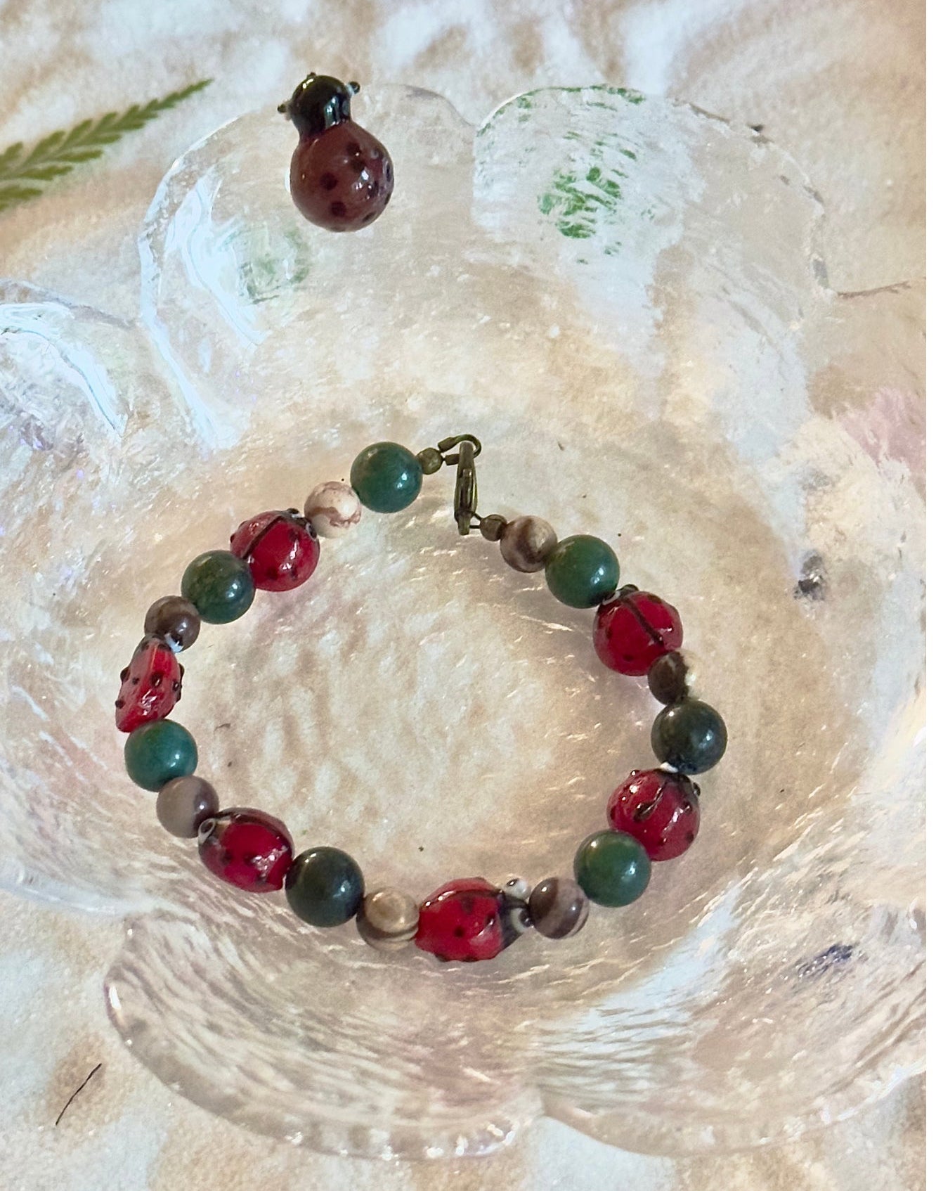 Handmade red and green beaded ladybug bracelet photographed in a textured glass dish.