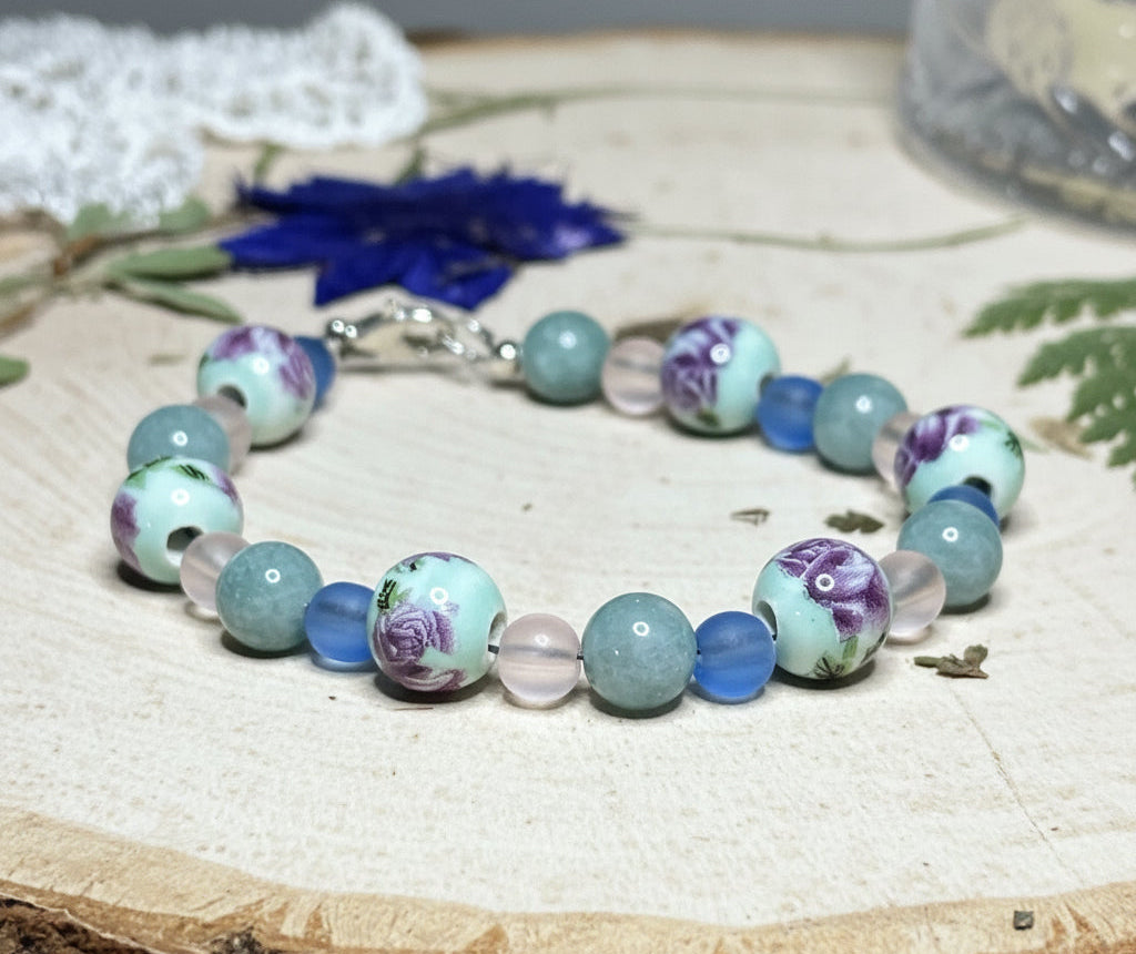 Handmade lavender coastal bracelet with floral porcelain beads and frosted blue glass displayed on a rustic wood slice