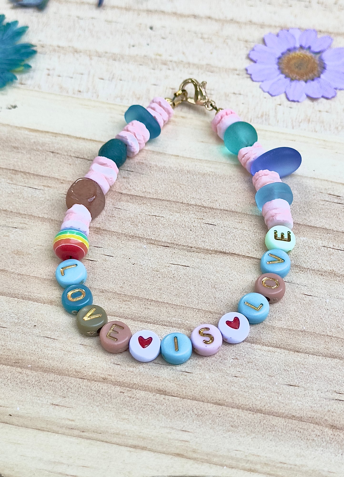 Flat lay of a handmade beaded bracelet spelling “LOVE IS LOVE,” featuring pastel beads, heart accents, sea glass details, and a gold clasp on a light wood background.