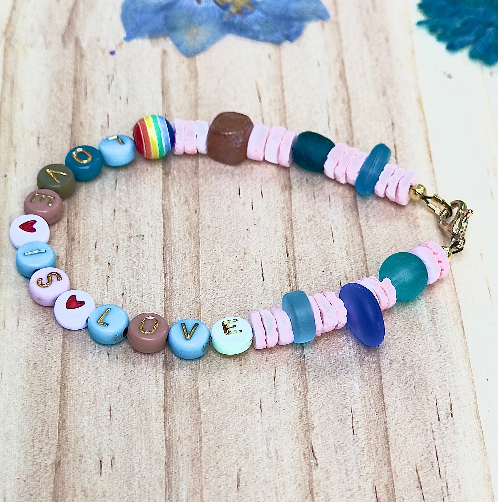 Angled flat lay of a pastel beaded “LOVE IS LOVE” bracelet showing sea glass beads, soft pink spacers, and gold-letter accents.