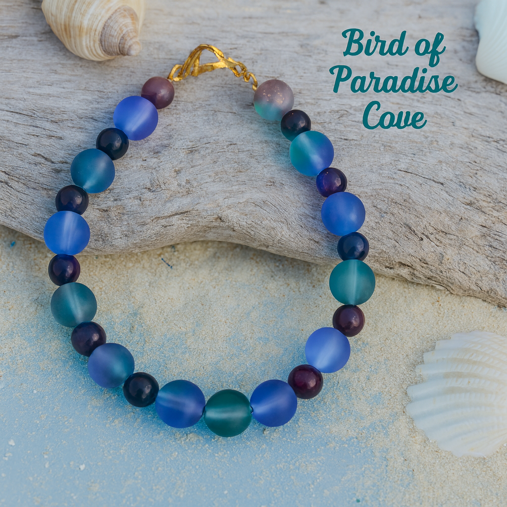 Lunar Twilight Galaxy handmade bracelet displayed on seashells with Bird of Paradise Cove branding, highlighting celestial-inspired blue, teal, purple, gray, and black beads.