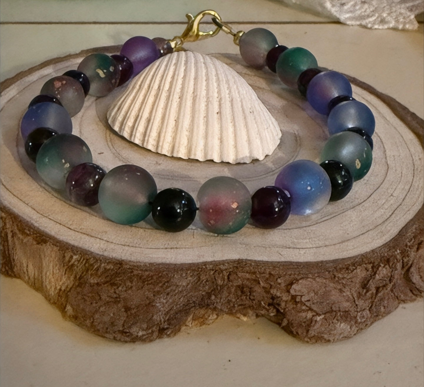 Lunar Twilight Galaxy celestial bracelet displayed on a natural wood slice with a seashell, featuring calming blue, teal, purple, gray, and black gemstone beads.