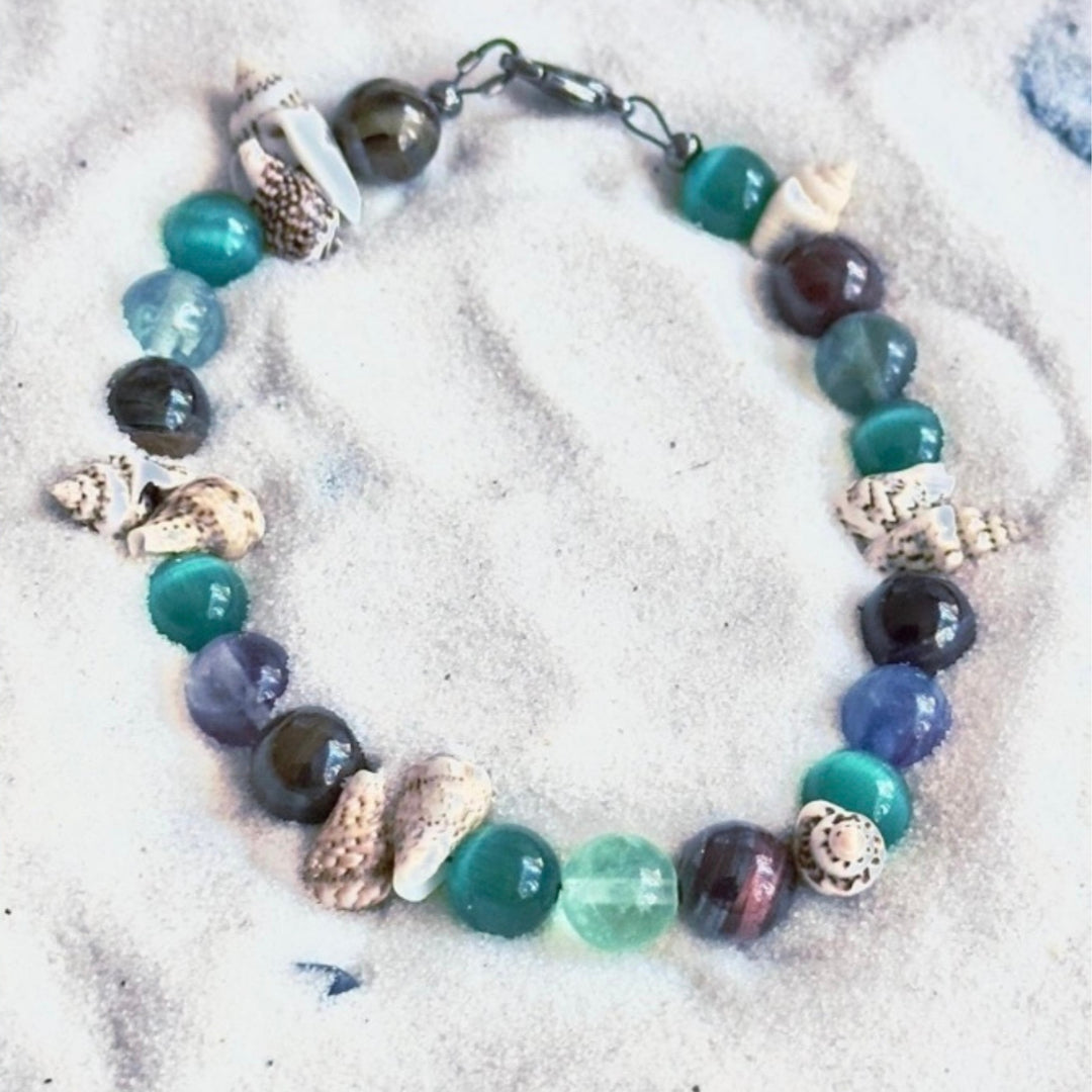 Ocean’s Treasure coastal bracelet photographed on white sand with shell accents and ocean-colored gemstone beads.