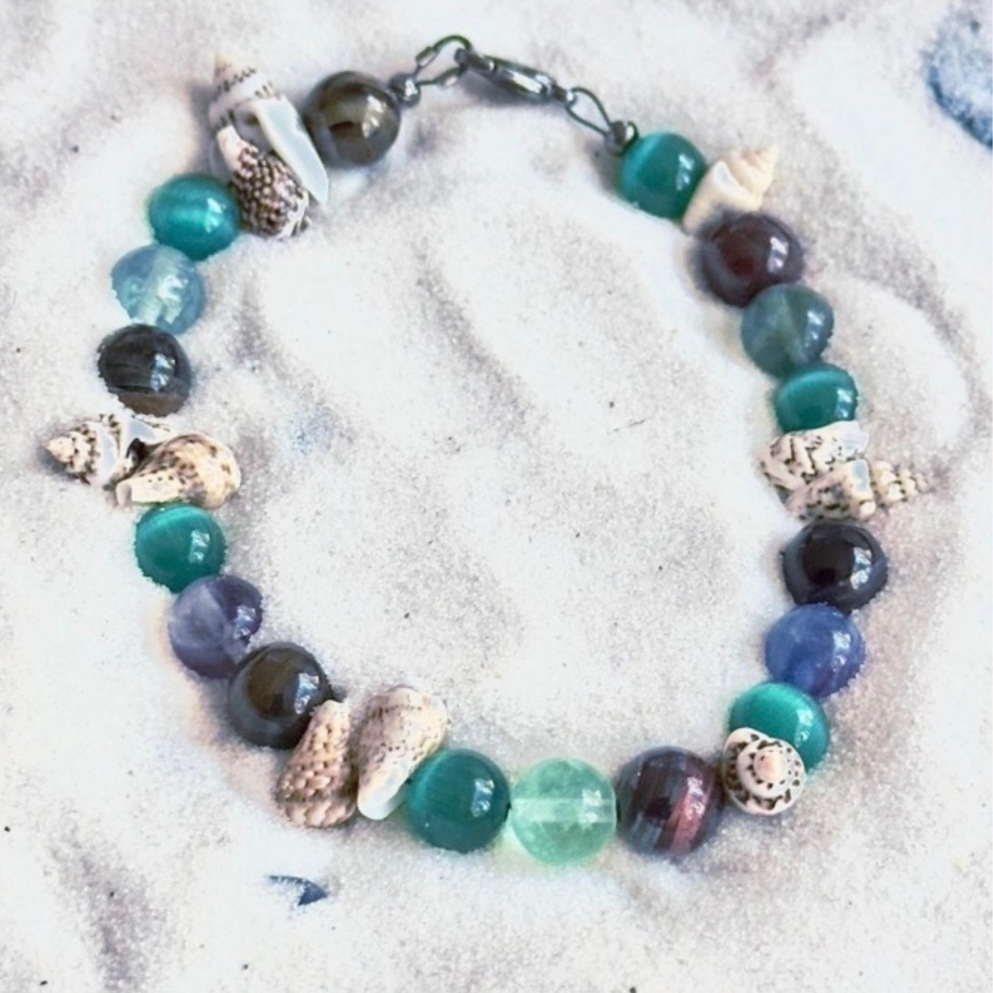 Ocean’s Treasure coastal bracelet photographed on white sand with shell accents and ocean-colored gemstone beads.