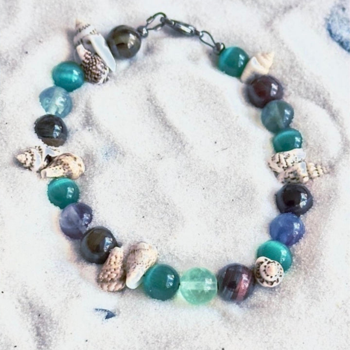 Ocean’s Treasure coastal bracelet photographed on white sand with shell accents and ocean-colored gemstone beads.