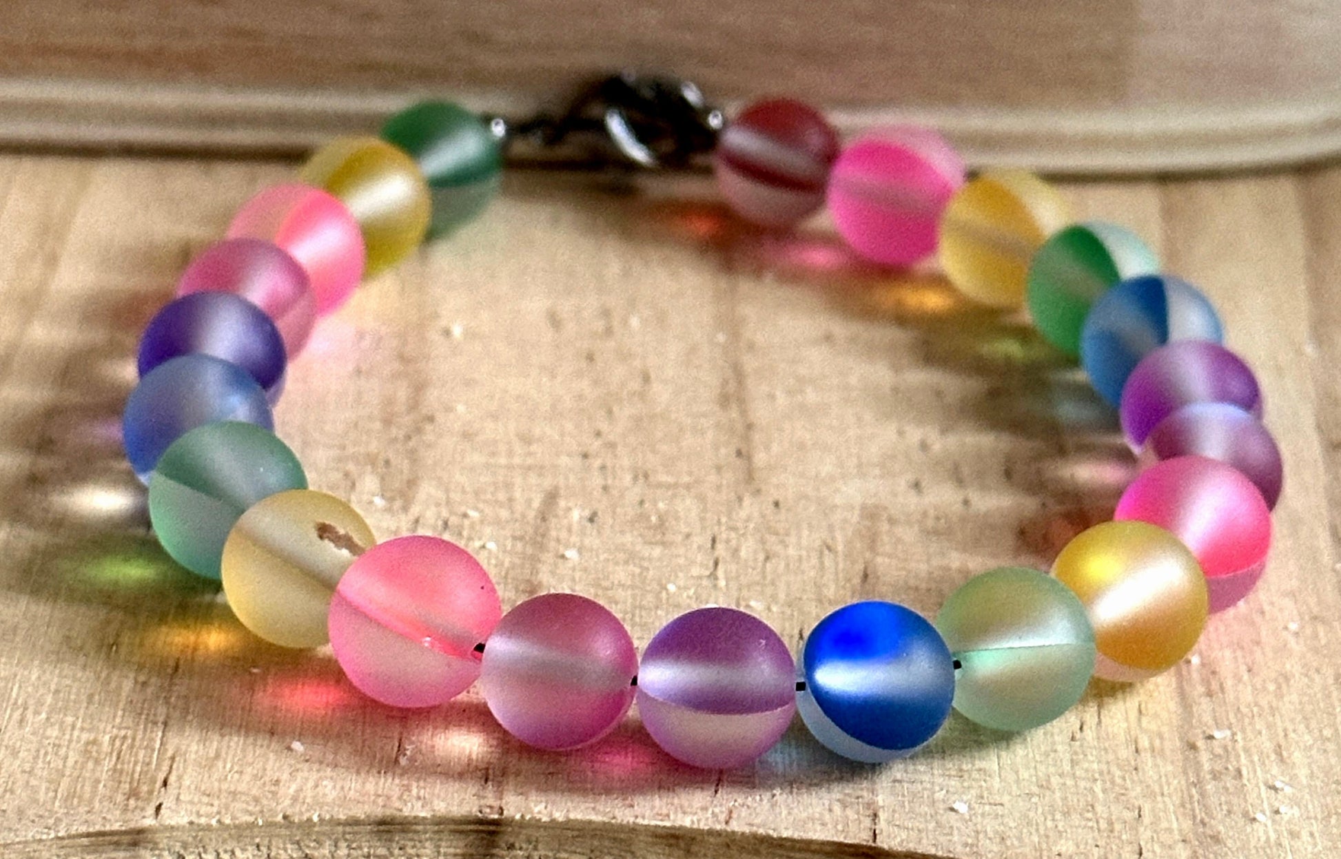 Handmade rainbow bracelet photographed on natural wood showing colorful frosted glass beads and lobster clasp closure.