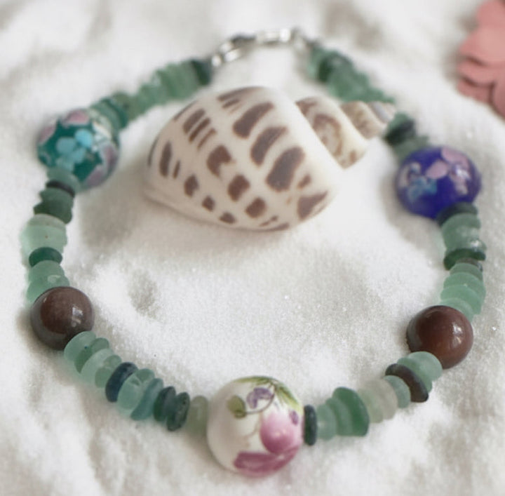 Sea glass floral bracelet displayed on sand with seashells and pastel flowers in coastal setting