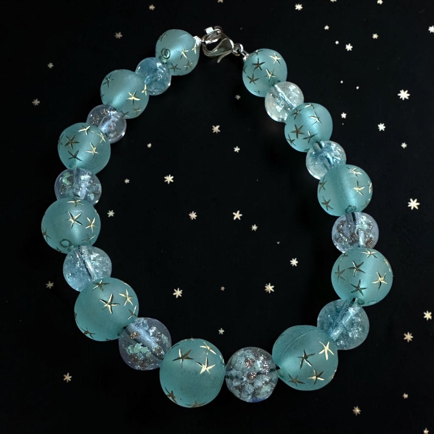 Celestial bracelet with aqua-blue star beads and crystal accents displayed against a starry black background inspired by the night sky.