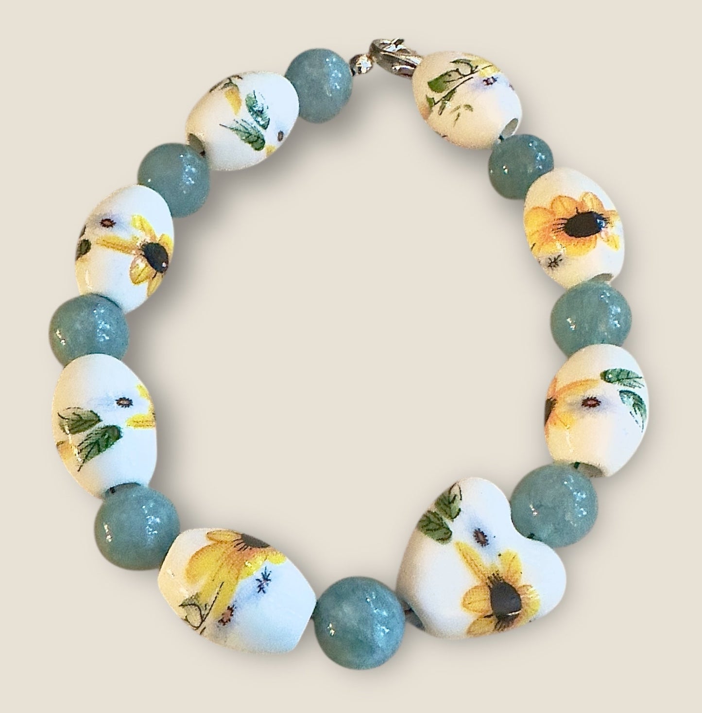 Handmade sunflower bracelet featuring floral porcelain beads and aqua glass beads styled on a rustic natural wood background with greenery accents.