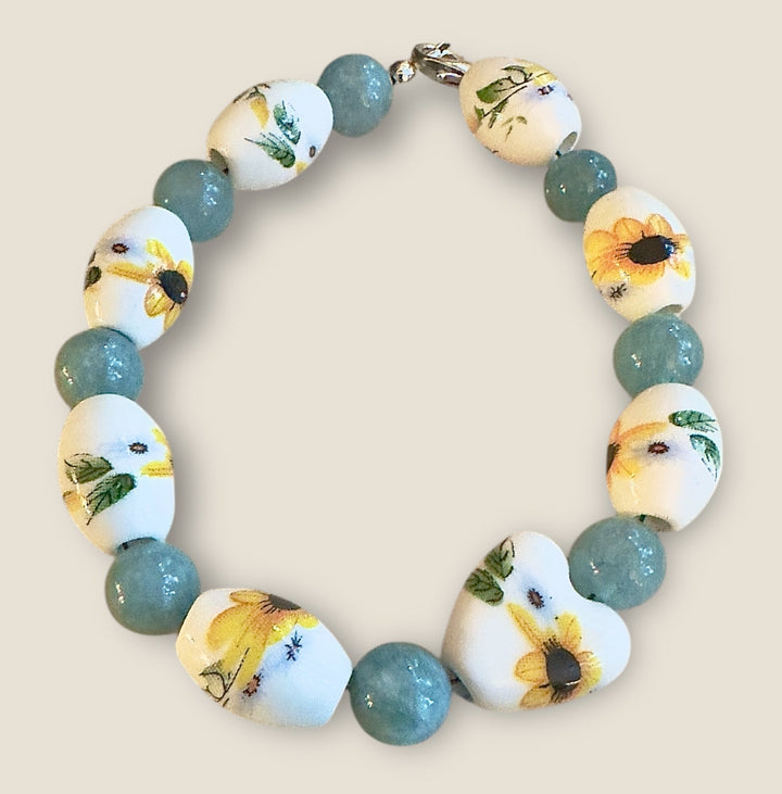 Handmade sunflower bracelet featuring floral porcelain beads and aqua glass beads styled on a rustic natural wood background with greenery accents.
