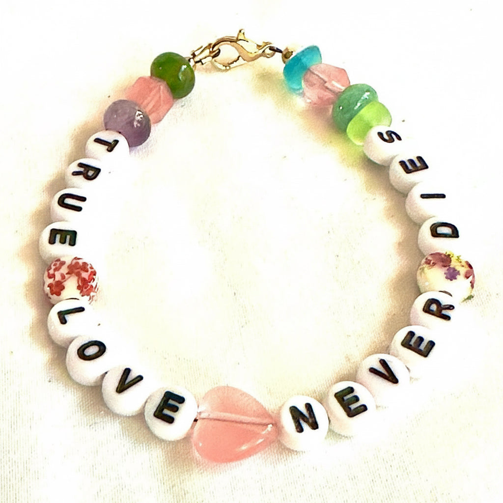 Handmade True Love Never Dies bracelet featuring white letter beads, pastel pink, green, purple, and turquoise accent beads, floral ceramic beads, a pink heart centerpiece, and a gold clasp on a light background.