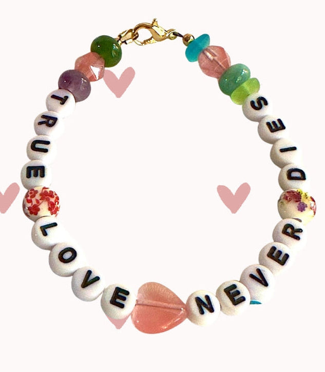 True Love Never Dies bracelet displayed on a soft pink background with scattered heart illustrations, highlighting the bracelet’s romantic and meaningful design.