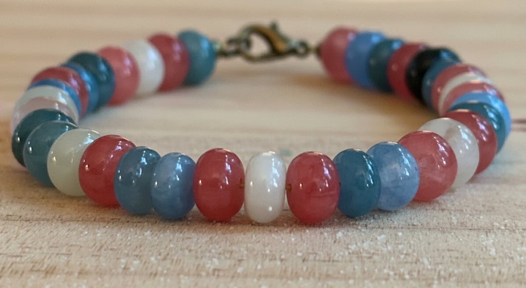 Pastel pink, blue, and white beaded Pride bracelet photographed on a rustic wood surface.