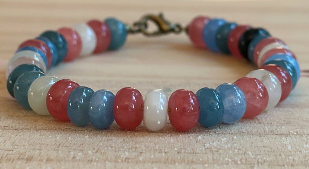 Pastel pink, blue, and white beaded Pride bracelet photographed on a rustic wood surface.