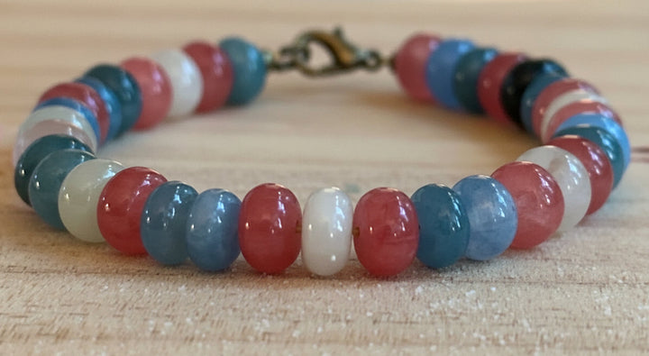 Pastel pink, blue, and white beaded Pride bracelet photographed on a rustic wood surface.