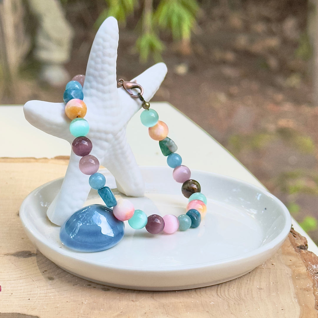 Colorful Earth and Sea bracelet displayed outdoors on a small white jewelry stand with soft natural scenery in the background.