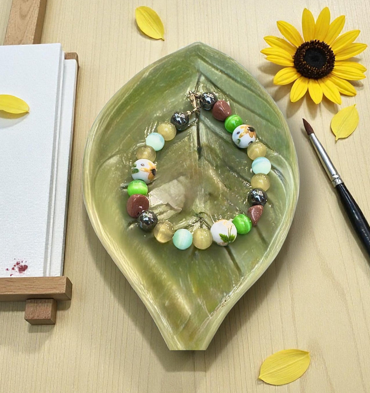 Willow Garden handmade bracelet arranged in a green leaf-shaped dish beside a sunflower, notebook, and pen on a pale green surface.