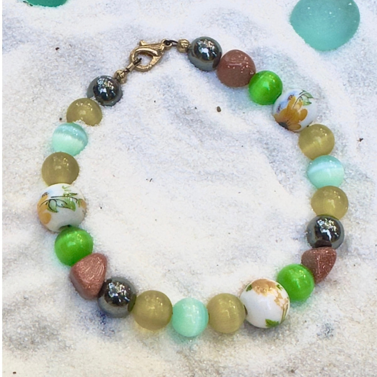 Willow Garden handmade bracelet arranged on sand background with aqua accents, displaying floral-patterned beads, glossy green rounds, and earthy golden tones.